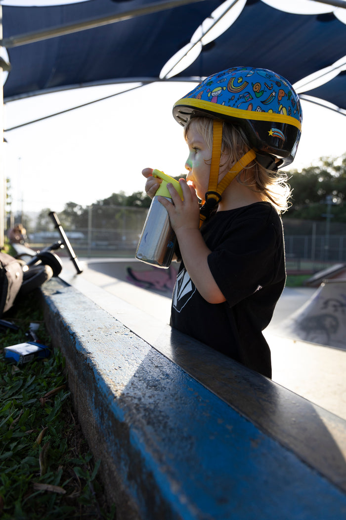 A child wearing a blue and yellow patterned skate helmet and a black t-shirt stands at a skatepark, drinking from a metal cup under a blue canopy.