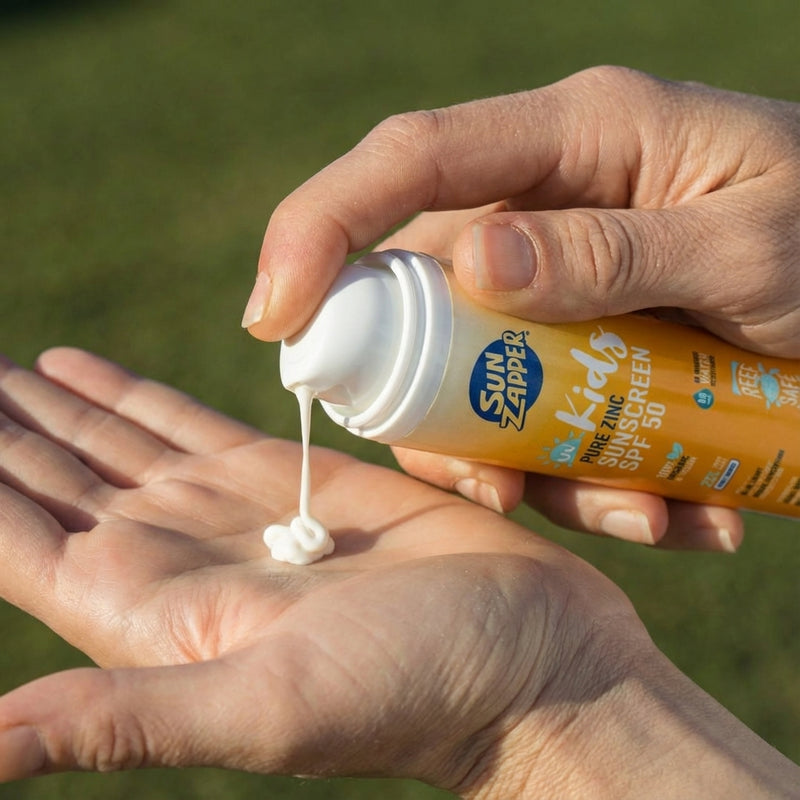 Hand holding a bottle of Sun Zapper Kids Pure & Zinc sunscreen with cream being dispensed onto another hand against a grassy background.