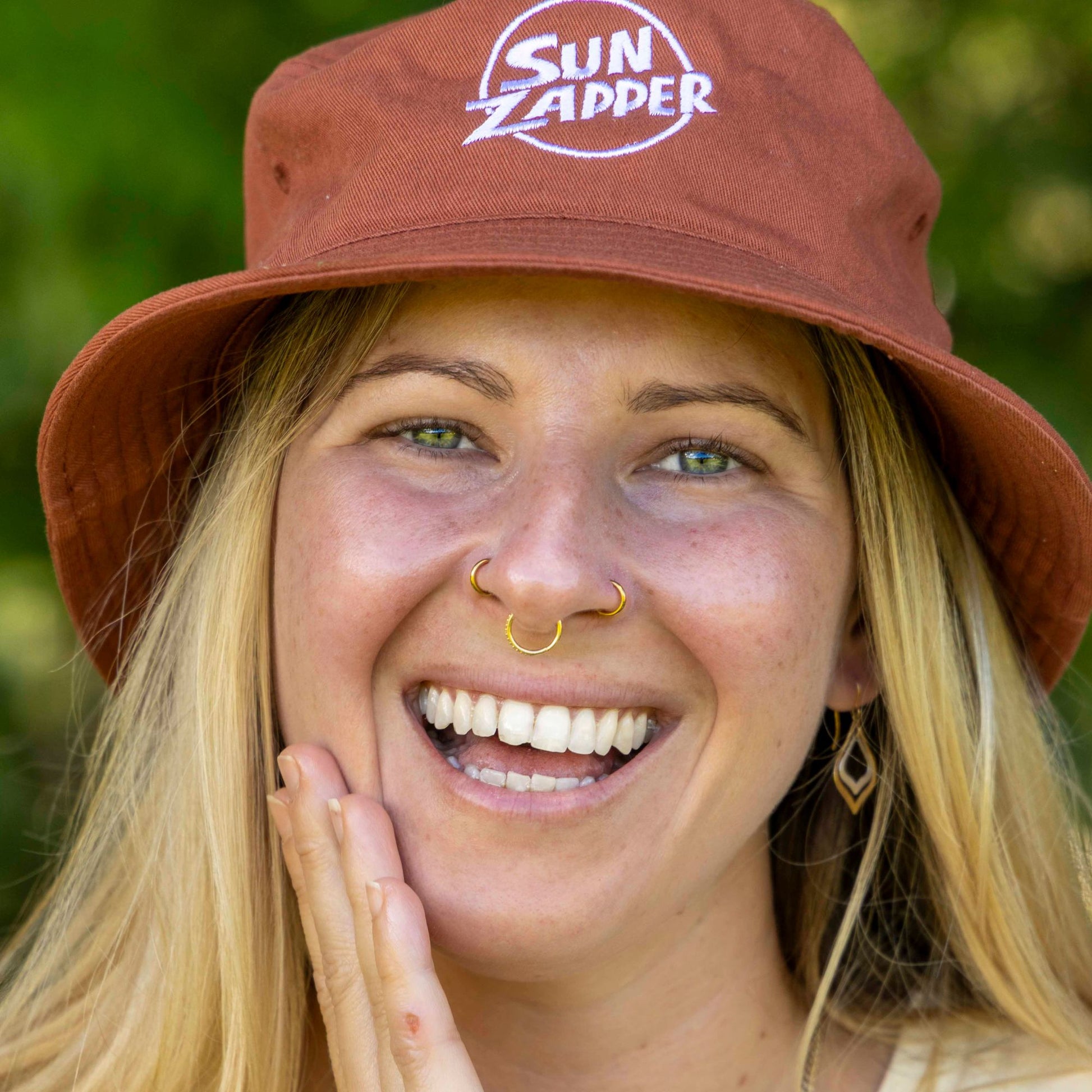 Person wearing a brown 'Sun Zapper' hat with a blurred green background applying face sunscreen